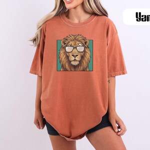 May include: A rust-colored, short-sleeved t-shirt featuring a graphic print of a lion wearing glasses centered on the chest. The lion is depicted within a teal square frame. The word "Yam" is printed in black text in the upper right corner.