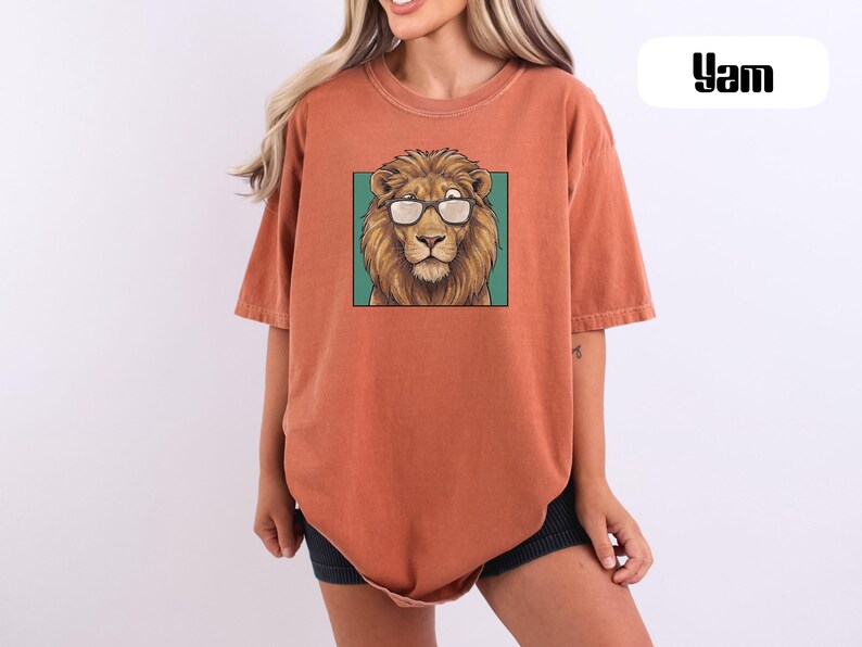 May include: A rust-colored, short-sleeved t-shirt featuring a graphic print of a lion wearing glasses centered on the chest. The lion is depicted within a teal square frame. The word "Yam" is printed in black text in the upper right corner.