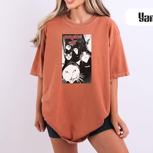 May include: A rust-colored short-sleeve t-shirt featuring a central black and white graphic print of multiple cats wearing hoodies with the text "gangster cat" in red lettering. The shirt has a crew neckline and a loose, relaxed fit throughout.