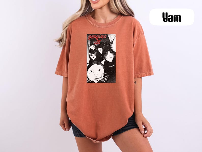 May include: A rust-colored short-sleeve t-shirt featuring a central black and white graphic print of multiple cats wearing hoodies with the text "gangster cat" in red lettering. The shirt has a crew neckline and a loose, relaxed fit throughout.