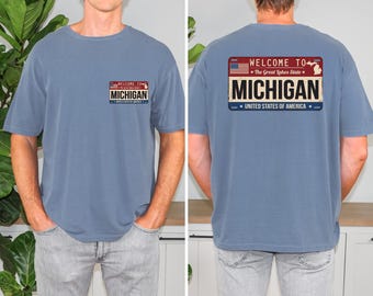 May include: A blue t-shirt featuring a graphic on the front left chest and a larger version on the back. The graphic resembles a license plate with the text "WELCOME TO The Great Lakes State", "MICHIGAN", and "UNITED STATES OF AMERICA".
