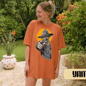 May include: An orange short-sleeve t-shirt featuring a graphic print of a scarecrow wearing a hat against a yellow circular background. The scarecrow is pointing forward with its right hand. The shirt has a crew neckline and a loose fit.