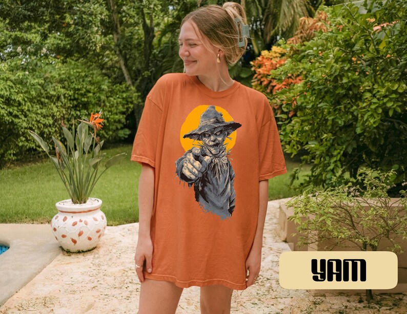 May include: An orange short-sleeve t-shirt featuring a graphic print of a scarecrow wearing a hat against a yellow circular background. The scarecrow is pointing forward with its right hand. The shirt has a crew neckline and a loose fit.