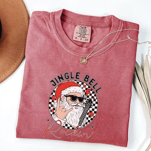 May include: A dusty rose short-sleeve t-shirt featuring a graphic of Santa Claus wearing sunglasses, making a rock-and-roll hand gesture, and holding an electric guitar. The text reads JINGLE BELL Rockin' inside a black and white checkered circle.