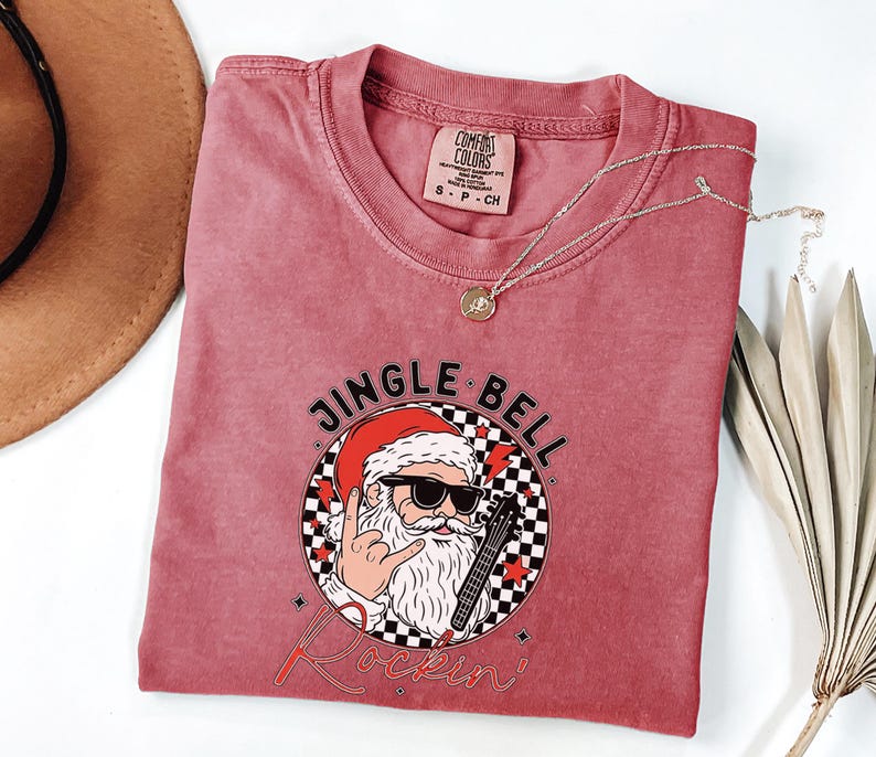 May include: A dusty rose short-sleeve t-shirt featuring a graphic of Santa Claus wearing sunglasses, making a rock-and-roll hand gesture, and holding an electric guitar. The text reads JINGLE BELL Rockin' inside a black and white checkered circle.