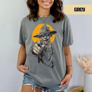 May include: A grey short-sleeve t-shirt featuring a graphic print of a scarecrow wearing a hat, pointing forward with a finger. The scarecrow is set against a solid yellow circular background. The word GREY is printed in a box at the top right.
