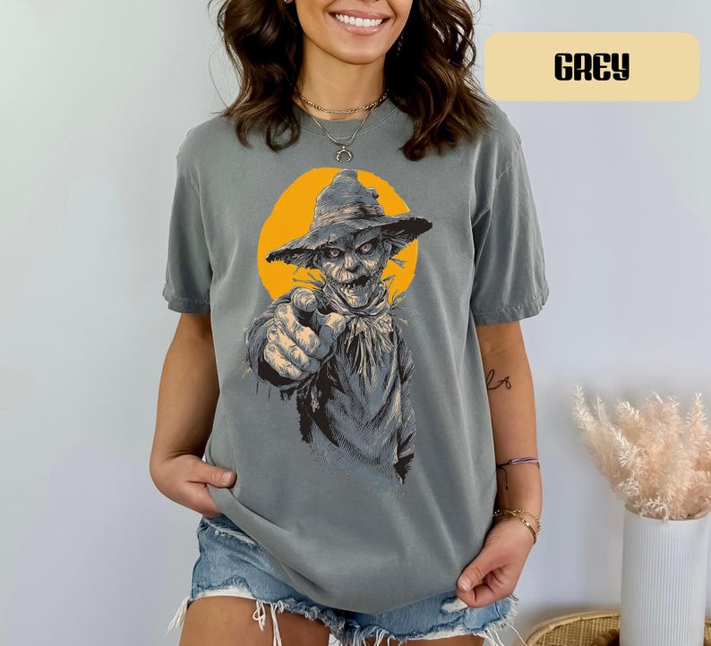 May include: A grey short-sleeve t-shirt featuring a graphic print of a scarecrow wearing a hat, pointing forward with a finger. The scarecrow is set against a solid yellow circular background. The word GREY is printed in a box at the top right.