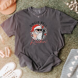 May include: A charcoal gray short-sleeve t-shirt featuring a graphic of Santa Claus wearing sunglasses and making a rock-and-roll hand gesture. The circular design includes a black and white checkered pattern, red hearts, and the text JINGLE BELL Rockin.
