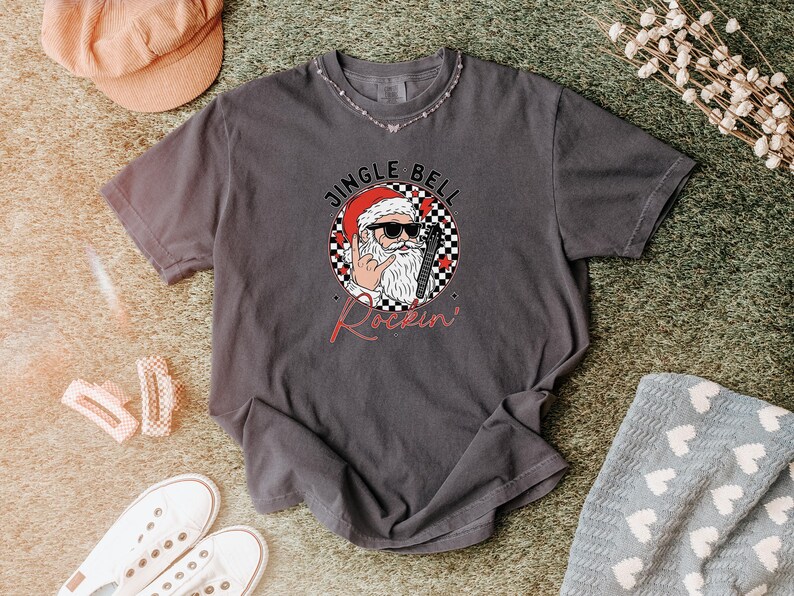 May include: A charcoal gray short-sleeve t-shirt featuring a graphic of Santa Claus wearing sunglasses and making a rock-and-roll hand gesture. The circular design includes a black and white checkered pattern, red hearts, and the text JINGLE BELL Rockin.