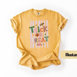 May include: A mustard yellow short-sleeve t-shirt featuring a graphic print with vertical light purple stripes. The text reads Trick Treat in colorful, patterned letters, accompanied by illustrations of a pumpkin and two candies.