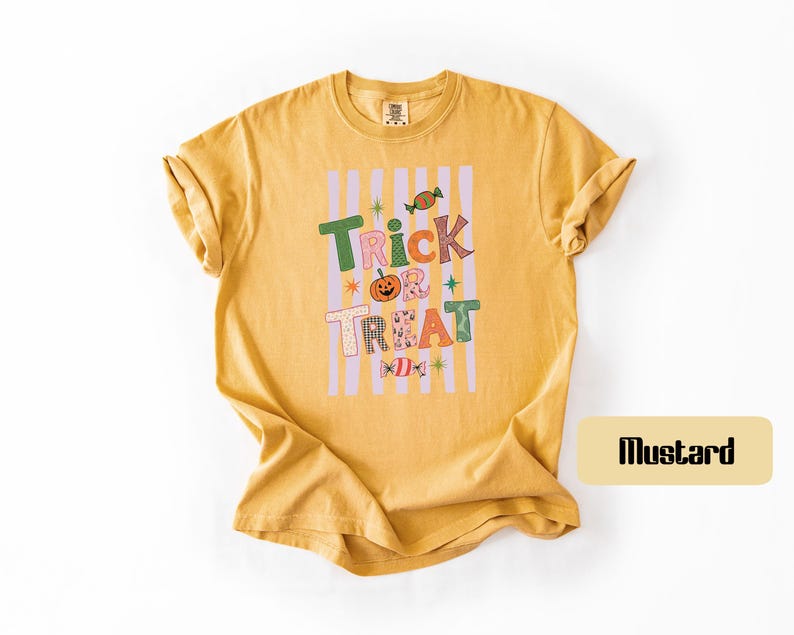 May include: A mustard yellow short-sleeve t-shirt featuring a graphic print with vertical light purple stripes. The text reads Trick Treat in colorful, patterned letters, accompanied by illustrations of a pumpkin and two candies.