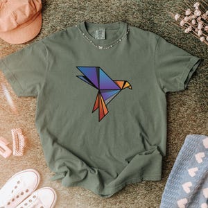 Puede incluir: Una camiseta de manga corta color verde oliva con un gr&aacute;fico de p&aacute;jaro de origami geom&eacute;trico en el pecho. El dise&ntilde;o del p&aacute;jaro est&aacute; compuesto por formas angulares en un degradado de colores azul, morado y naranja con bordes negros.