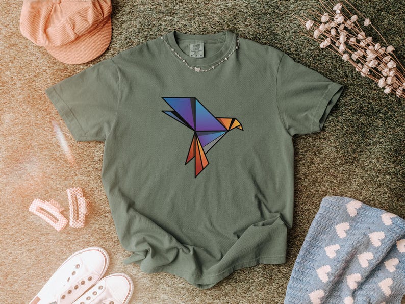 Puede incluir: Una camiseta de manga corta color verde oliva con un gr&aacute;fico de p&aacute;jaro de origami geom&eacute;trico en el pecho. El dise&ntilde;o del p&aacute;jaro est&aacute; compuesto por formas angulares en un degradado de colores azul, morado y naranja con bordes negros.