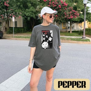 May include: A charcoal gray short-sleeve t-shirt featuring a black and white graphic print of multiple cats and the text "gangster cat" in red lettering. The shirt has a crew neckline and a relaxed fit, constructed from a solid-colored cotton fabric.