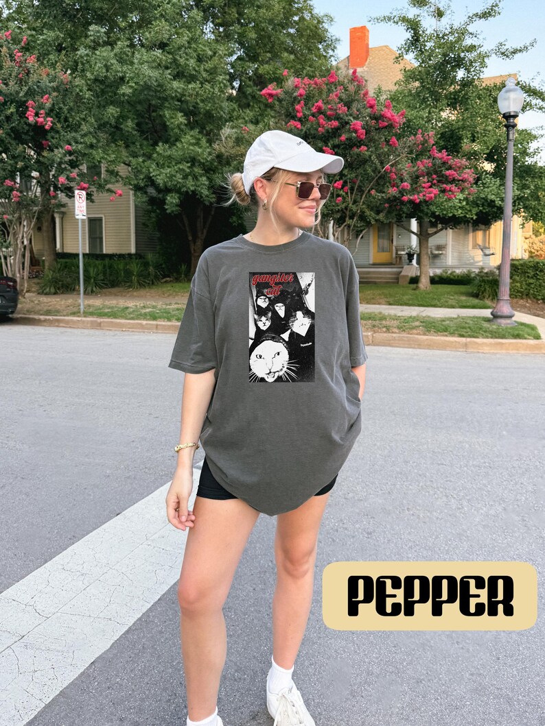 May include: A charcoal gray short-sleeve t-shirt featuring a black and white graphic print of multiple cats and the text "gangster cat" in red lettering. The shirt has a crew neckline and a relaxed fit, constructed from a solid-colored cotton fabric.