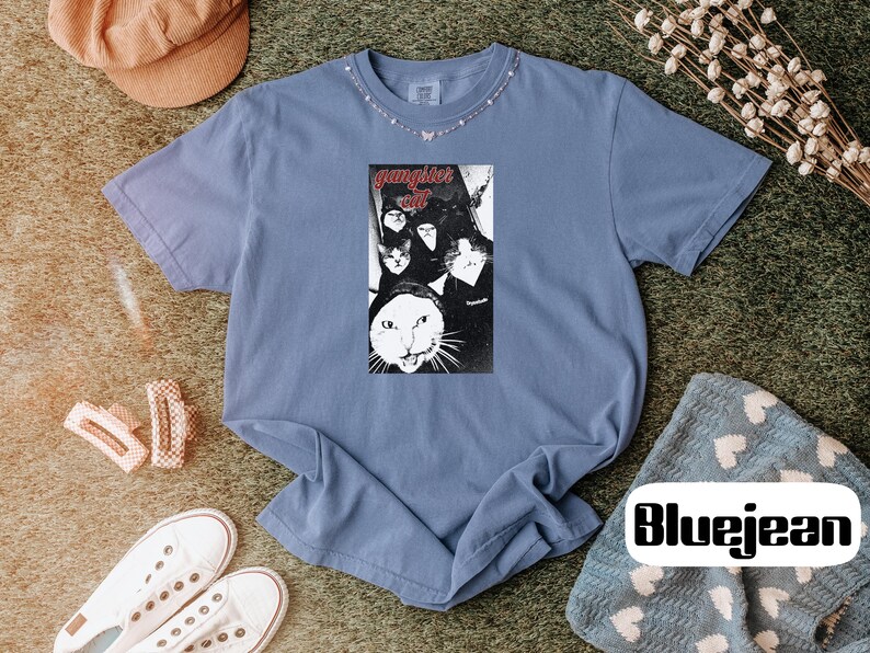 May include: A blue short-sleeve t-shirt featuring a central rectangular graphic print of multiple cats with the text "gangster cat" in red lettering. The shirt has a crew neckline and a relaxed fit, showcasing a vintage-inspired wash.