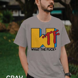May include: A gray short-sleeved t-shirt featuring a large yellow block letter W with a blue shadow and red stylized TF letters overlapping it. Below the graphic, the text WHAT THE FUCK is printed in black capital letters with a registered symbol.