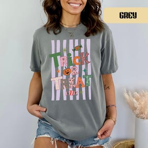 May include: A grey short-sleeve t-shirt featuring a graphic print with vertical light purple stripes and the text "TRICK OR TREAT" in colorful, patterned letters. The design includes a small orange pumpkin and candy illustrations.