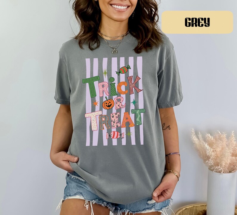 May include: A grey short-sleeve t-shirt featuring a graphic print with vertical light purple stripes and the text "TRICK OR TREAT" in colorful, patterned letters. The design includes a small orange pumpkin and candy illustrations.