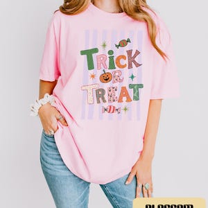 May include: A light pink short-sleeved t-shirt featuring a graphic print with the text Trick or Treat in various colors and patterns. The design includes a central orange pumpkin, two candy illustrations, and small star accents over vertical light purple stripes.
