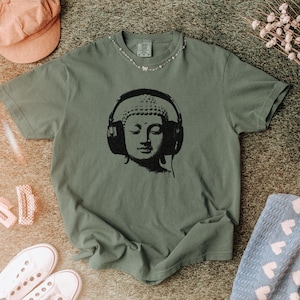 May include: A sage green short-sleeve t-shirt featuring a black graphic print of a Buddha head wearing large over-ear headphones. The shirt has a crew neckline and a relaxed fit, showcasing a detailed illustration centered on the chest area.