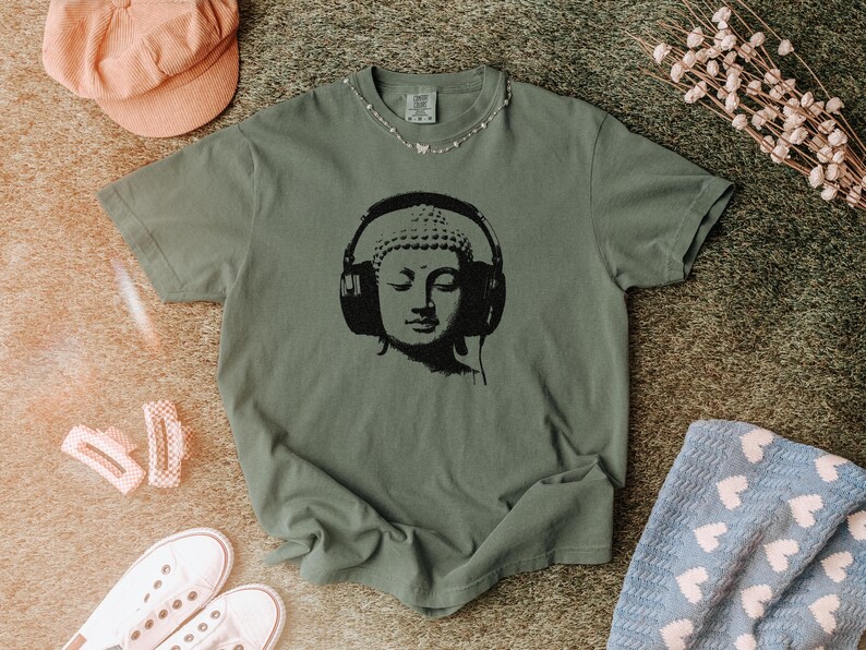 May include: A sage green short-sleeve t-shirt featuring a black graphic print of a Buddha head wearing large over-ear headphones. The shirt has a crew neckline and a relaxed fit, showcasing a detailed illustration centered on the chest area.