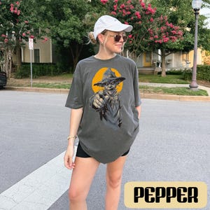 May include: A charcoal gray short-sleeve t-shirt featuring a graphic print of a scarecrow wearing a hat and pointing forward against a yellow circular background. The shirt is labeled with the color name Pepper in a rectangular box.