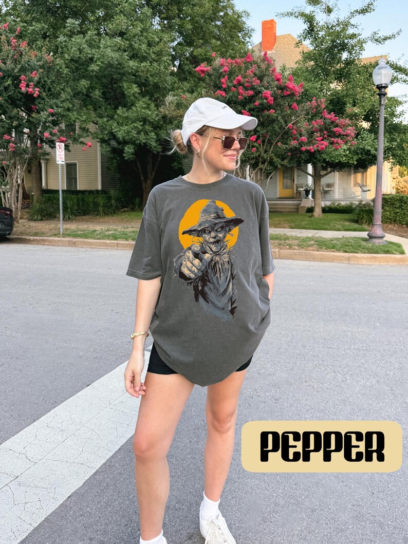 May include: A charcoal gray short-sleeve t-shirt featuring a graphic print of a scarecrow wearing a hat and pointing forward against a yellow circular background. The shirt is labeled with the color name Pepper in a rectangular box.