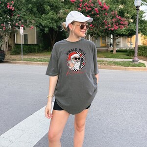 May include: A charcoal gray short-sleeve t-shirt featuring a graphic of Santa Claus wearing sunglasses and making a rock-and-roll hand gesture. The design includes a black and white checkered circle, red accents, and the text JINGLE BELL ROCKIN.