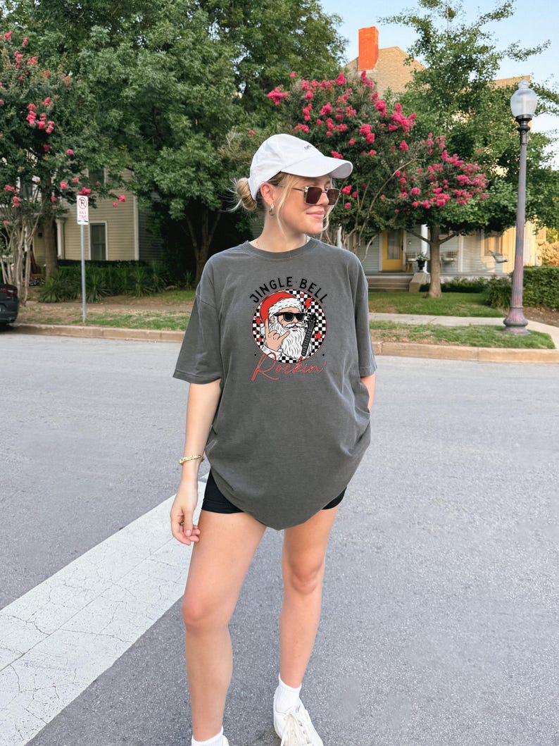 May include: A charcoal gray short-sleeve t-shirt featuring a graphic of Santa Claus wearing sunglasses and making a rock-and-roll hand gesture. The design includes a black and white checkered circle, red accents, and the text JINGLE BELL ROCKIN.