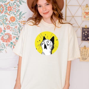May include: An off-white short-sleeve t-shirt featuring a circular graphic on the chest. The graphic displays a black and white line-art illustration of a cat with wide, surprised eyes and an open mouth, set against a solid bright yellow background.