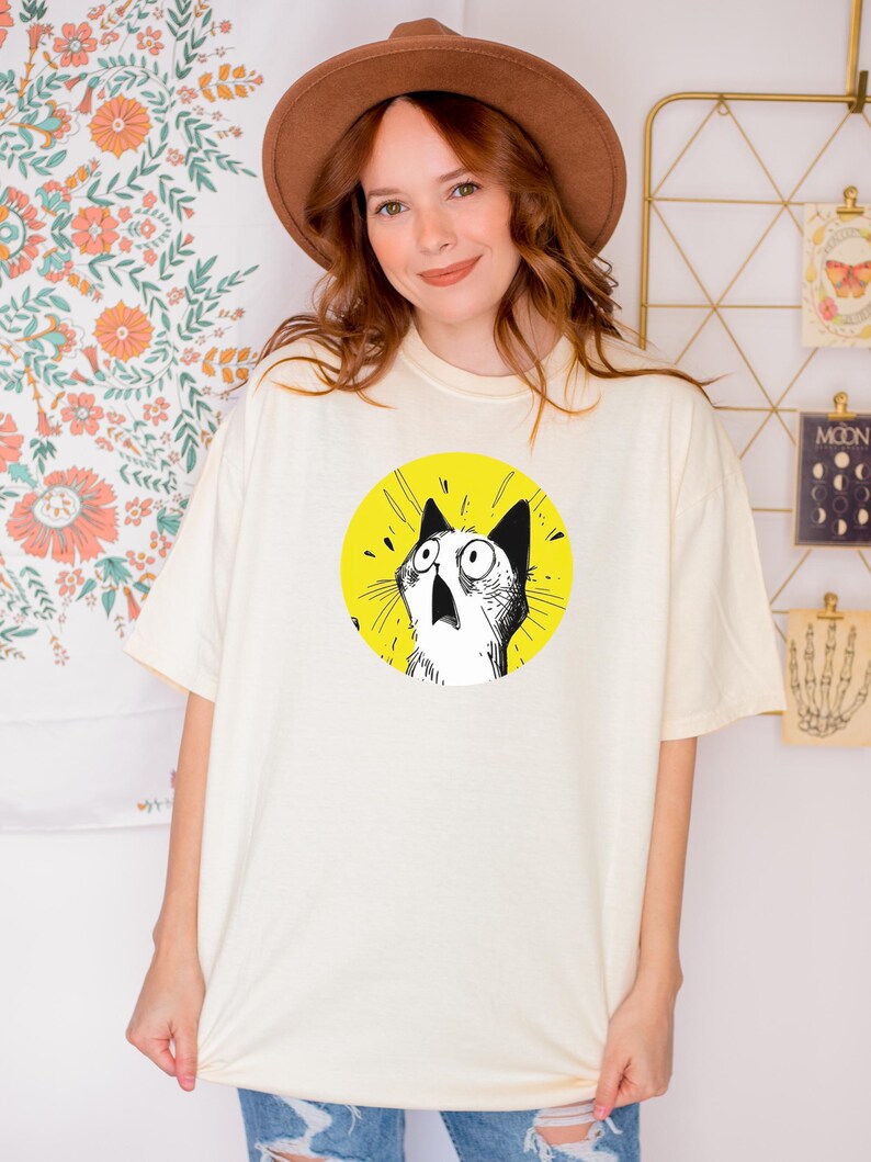 May include: An off-white short-sleeve t-shirt featuring a circular graphic on the chest. The graphic displays a black and white line-art illustration of a cat with wide, surprised eyes and an open mouth, set against a solid bright yellow background.