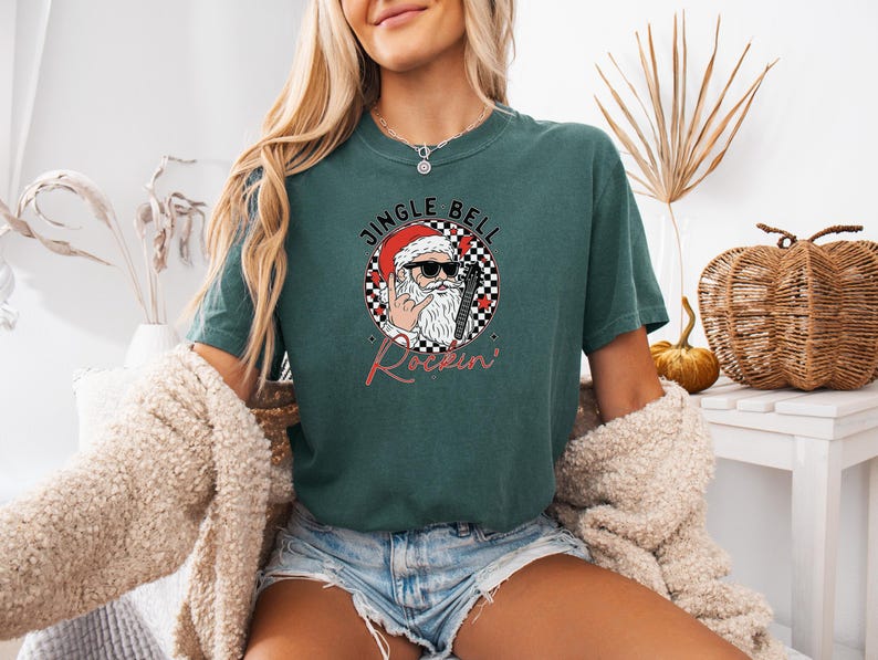 May include: A dark green short-sleeve t-shirt featuring a graphic of Santa Claus wearing sunglasses and making a rock-and-roll hand gesture. The design includes a black and white checkered circle, red accents, and the text JINGLE BELL Rockin'.