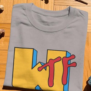 May include: A gray crew neck t-shirt featuring a large graphic print on the front. The design displays the letters KTF in a bold, blocky yellow font with blue 3D-style shading and a red, dripping-style font layered over the yellow letters.