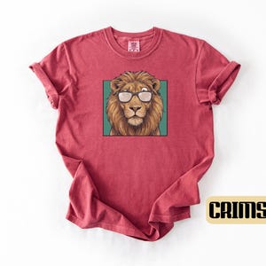May include: A crimson short-sleeve t-shirt featuring a central graphic of a brown lion wearing black-rimmed glasses. The lion is framed within a teal square. The fabric has a slightly textured, washed appearance with a crew neckline.