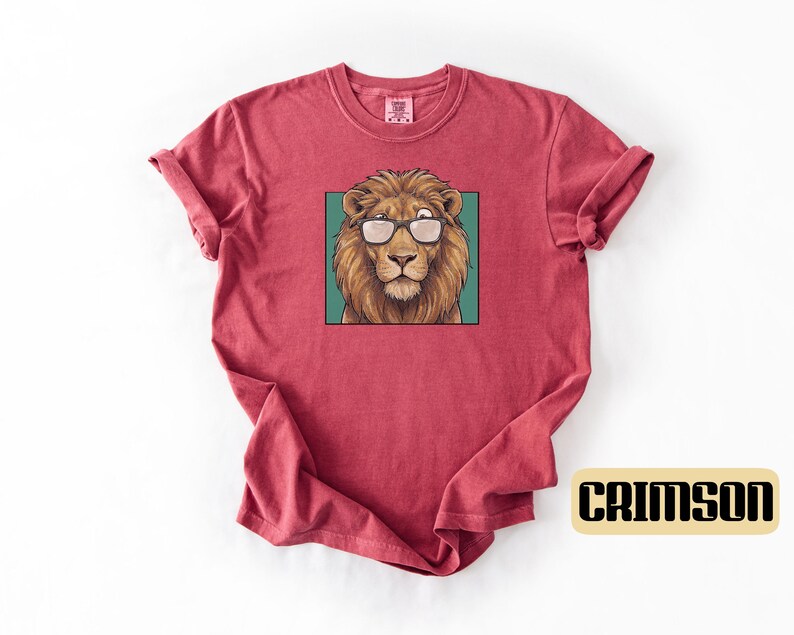 May include: A crimson short-sleeve t-shirt featuring a central graphic of a brown lion wearing black-rimmed glasses. The lion is framed within a teal square. The fabric has a slightly textured, washed appearance with a crew neckline.