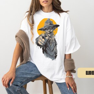 May include: A white t-shirt featuring a graphic print of a scarecrow wearing a hat and pointing forward. The scarecrow is rendered in shades of gray and brown, set against a solid yellow circular background. The word WHITE is printed in black.