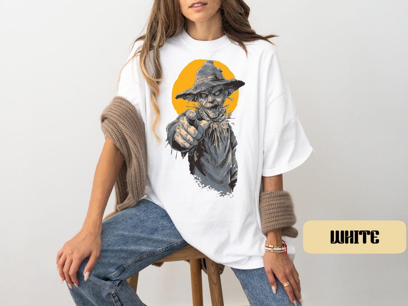 May include: A white t-shirt featuring a graphic print of a scarecrow wearing a hat and pointing forward. The scarecrow is rendered in shades of gray and brown, set against a solid yellow circular background. The word WHITE is printed in black.