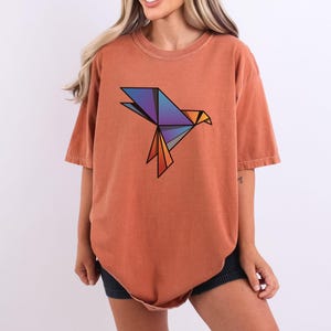 Puede incluir: Una camiseta de manga corta color terracota con un gr&aacute;fico de p&aacute;jaro de origami geom&eacute;trico en el pecho. El dise&ntilde;o del p&aacute;jaro est&aacute; compuesto por formas angulares en tonos azul, morado y naranja, delineadas en negro, creando una forma aviar.