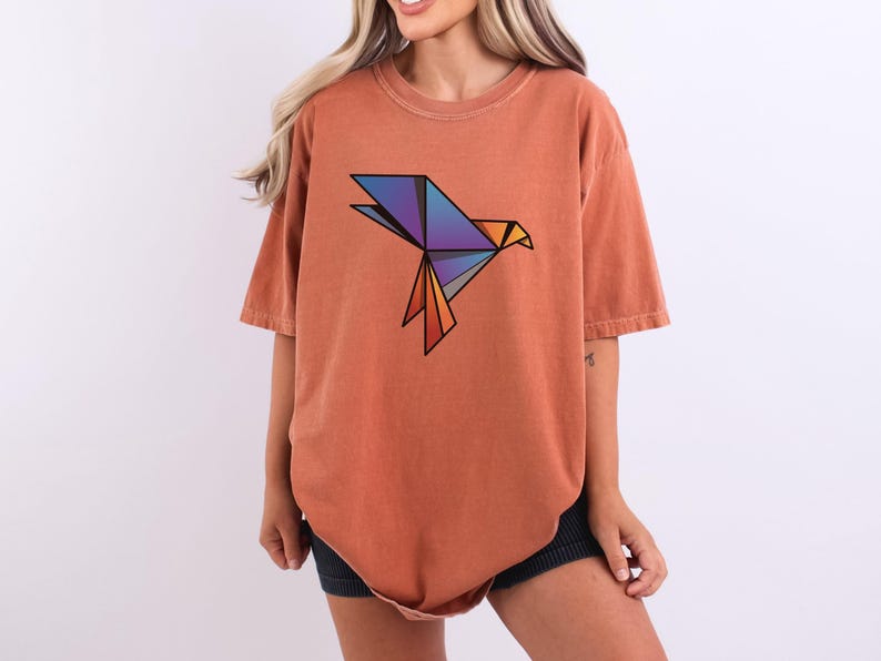 Puede incluir: Una camiseta de manga corta color terracota con un gr&aacute;fico de p&aacute;jaro de origami geom&eacute;trico en el pecho. El dise&ntilde;o del p&aacute;jaro est&aacute; compuesto por formas angulares en tonos azul, morado y naranja, delineadas en negro, creando una forma aviar.