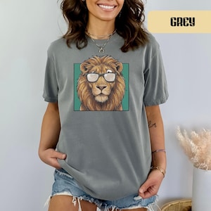 May include: A grey short-sleeved t-shirt featuring a graphic print of a lion wearing glasses. The lion is centered within a teal square frame. The shirt has a crew neckline and a relaxed fit, with the word GREY printed in a box at the top right.