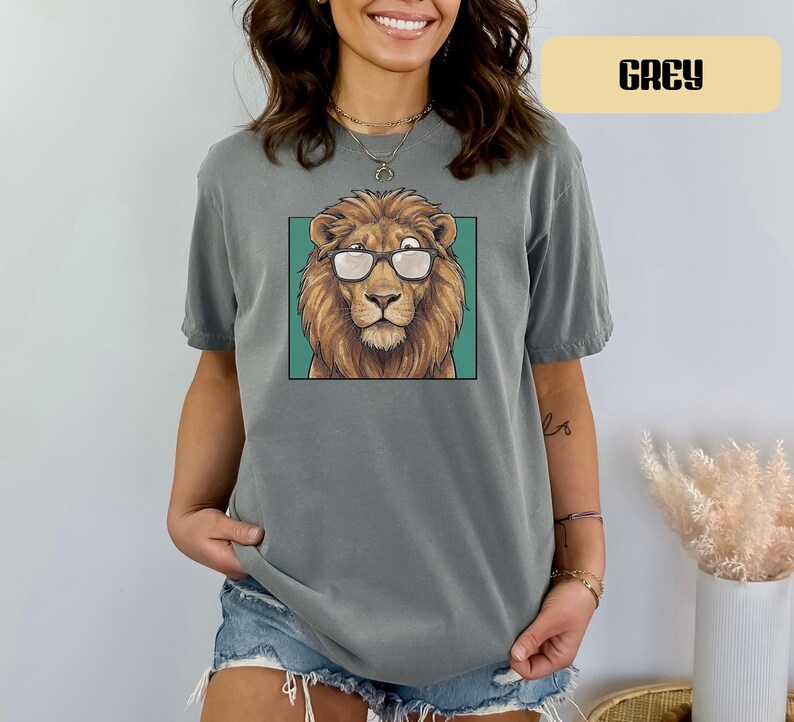 May include: A grey short-sleeved t-shirt featuring a graphic print of a lion wearing glasses. The lion is centered within a teal square frame. The shirt has a crew neckline and a relaxed fit, with the word GREY printed in a box at the top right.