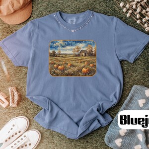 May include: A periwinkle blue short-sleeve t-shirt featuring a rectangular graphic print of a rural landscape with orange pumpkins in a field, a rustic barn, and a cloudy sky. The shirt has a crew neckline and a relaxed fit, showcasing a detailed scene.