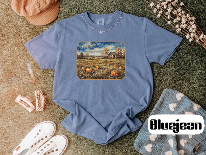 May include: A periwinkle blue short-sleeve t-shirt featuring a rectangular graphic print of a rural landscape with orange pumpkins in a field, a rustic barn, and a cloudy sky. The shirt has a crew neckline and a relaxed fit, showcasing a detailed scene.