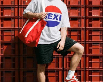 May include: A white short-sleeved t-shirt with a circular red and blue graphic featuring the text SEXI, paired with black shorts, a red canvas tote bag with white straps, red and white sneakers, white socks, and red-framed sunglasses.