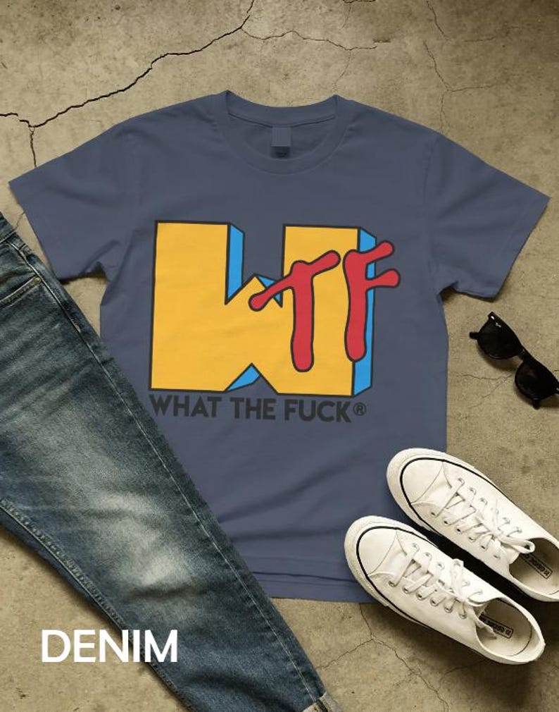 May include: A slate blue short-sleeve t-shirt featuring a large yellow block letter W with a red script TF overlay. Below the graphic, the text reads WHAT THE FUCK in black capital letters. The shirt is displayed alongside blue denim jeans.