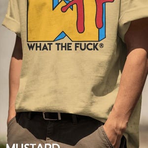 May include: A mustard yellow t-shirt featuring a large graphic print with the yellow block letters WTF, accented by a red script overlay and blue shadows. Below the graphic, the text WHAT THE FUCK is printed in black capital letters.