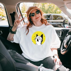 May include: A white short-sleeved t-shirt featuring a circular graphic of a surprised cat with wide eyes and an open mouth against a bright yellow background. The cat illustration is rendered in black and white ink style with radiating lines.
