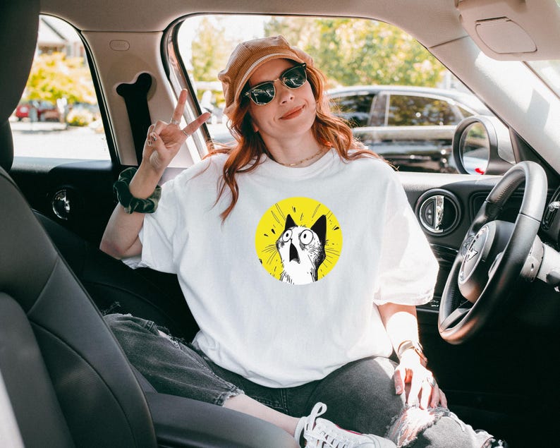 May include: A white short-sleeved t-shirt featuring a circular graphic of a surprised cat with wide eyes and an open mouth against a bright yellow background. The cat illustration is rendered in black and white ink style with radiating lines.