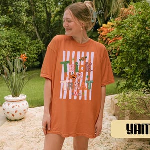 May include: An orange short-sleeved t-shirt featuring a graphic print with white vertical stripes and the text "TRICK TREAT" in colorful, patterned letters. The design includes small illustrations of a pumpkin, candy, and stars.